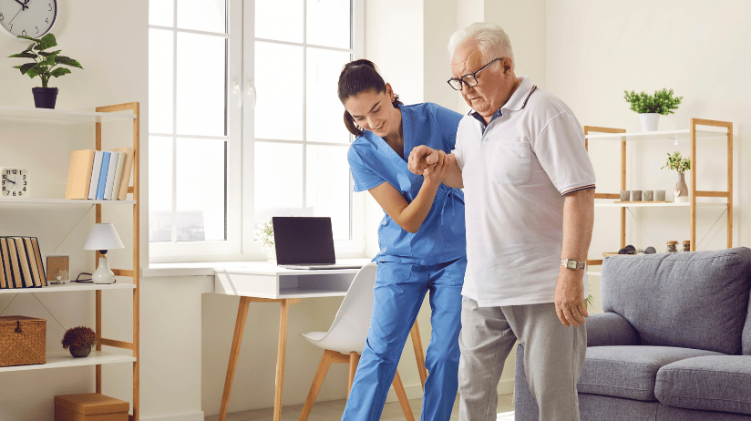 Female caregiver helping an elderly man walk in the living room