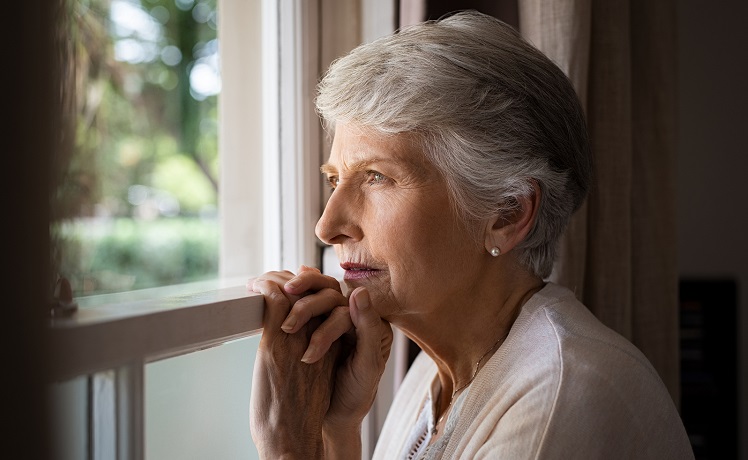 Depressed senior woman at home feeling sad. Elderly woman looks sadly outside the window. Depressed lonely lady standing alone and looking through the window.