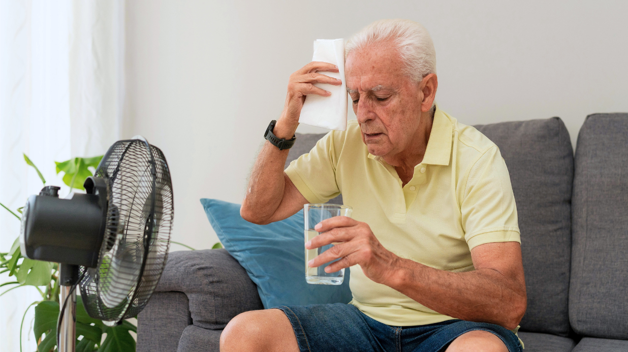 Senior man sitting on a couch while hot at home — holding a glass of water in front of a fan